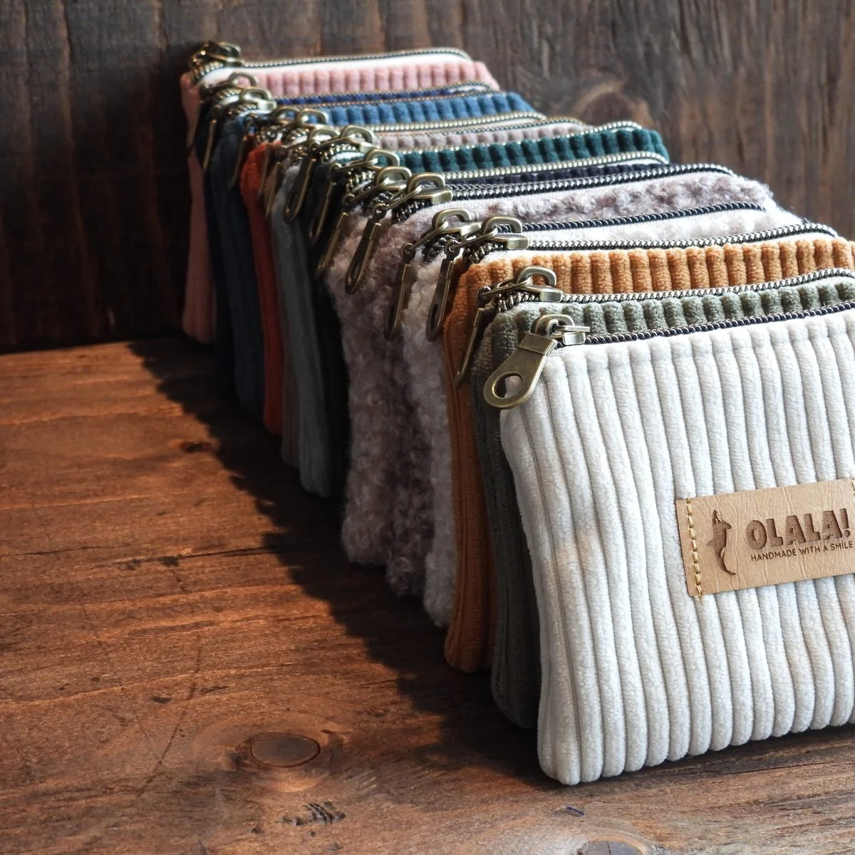 A row of zip pouches lined up on a wooden bench in corduroy and bouclé, from cream and sage through to rust and navy