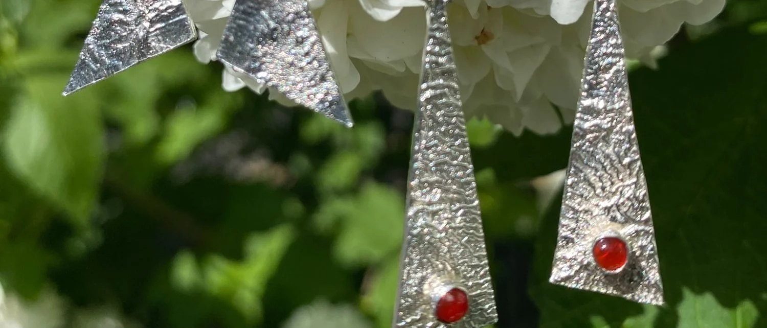 Four textured sterling silver triangle earrings hanging against a white hydrangea, the two longer pairs set with a small red carnelian near the base