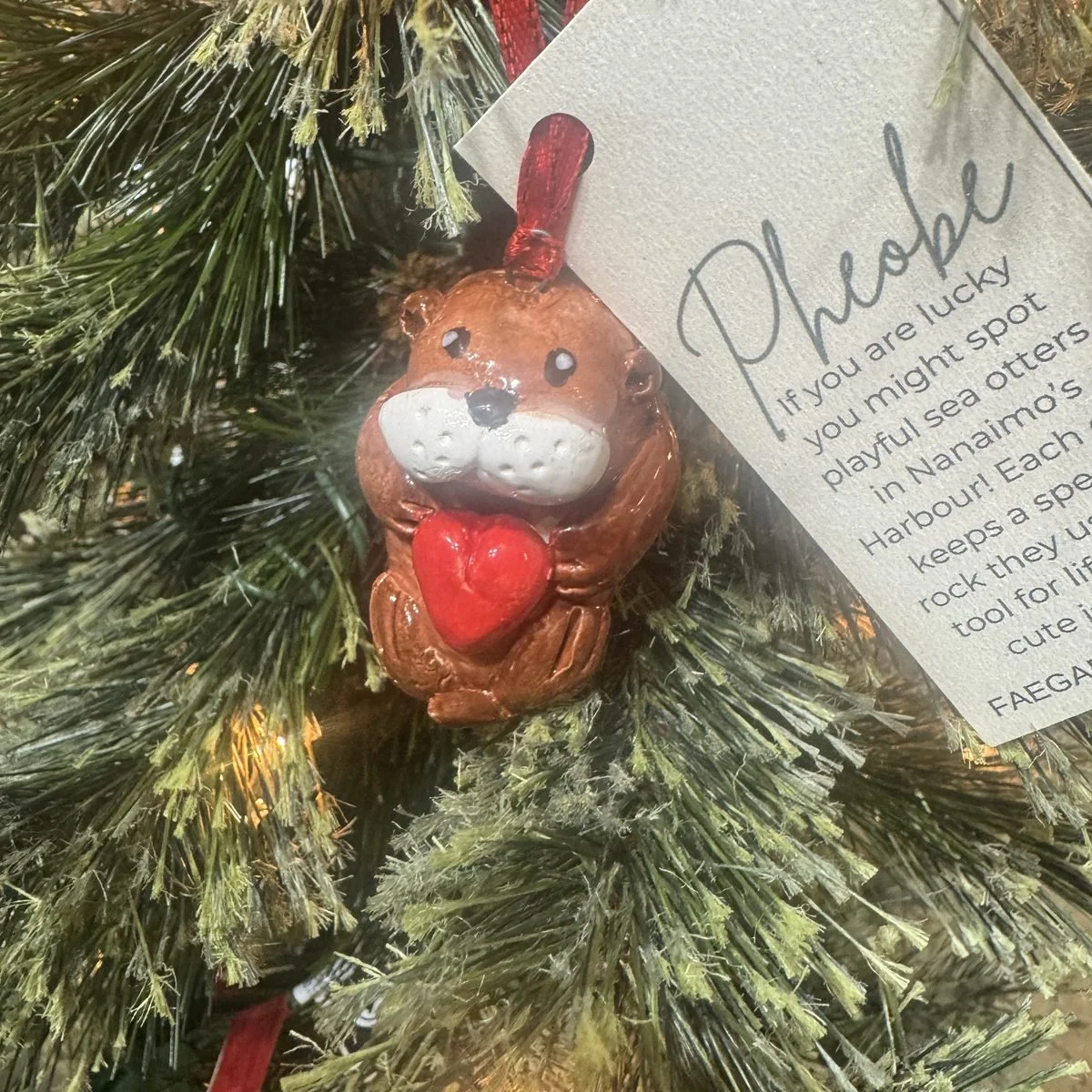A hand built sea otter ornament holding a red heart, hanging on a lit Christmas tree beside its card about sea otters in Nanaimo harbour