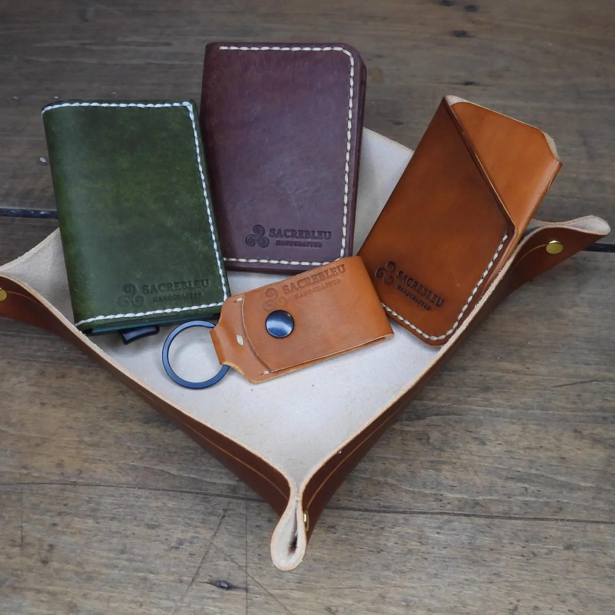 A leather valet tray holding a green and an oxblood wallet, a tan card holder and a keyring, each stamped Sacrebleu Handcrafted