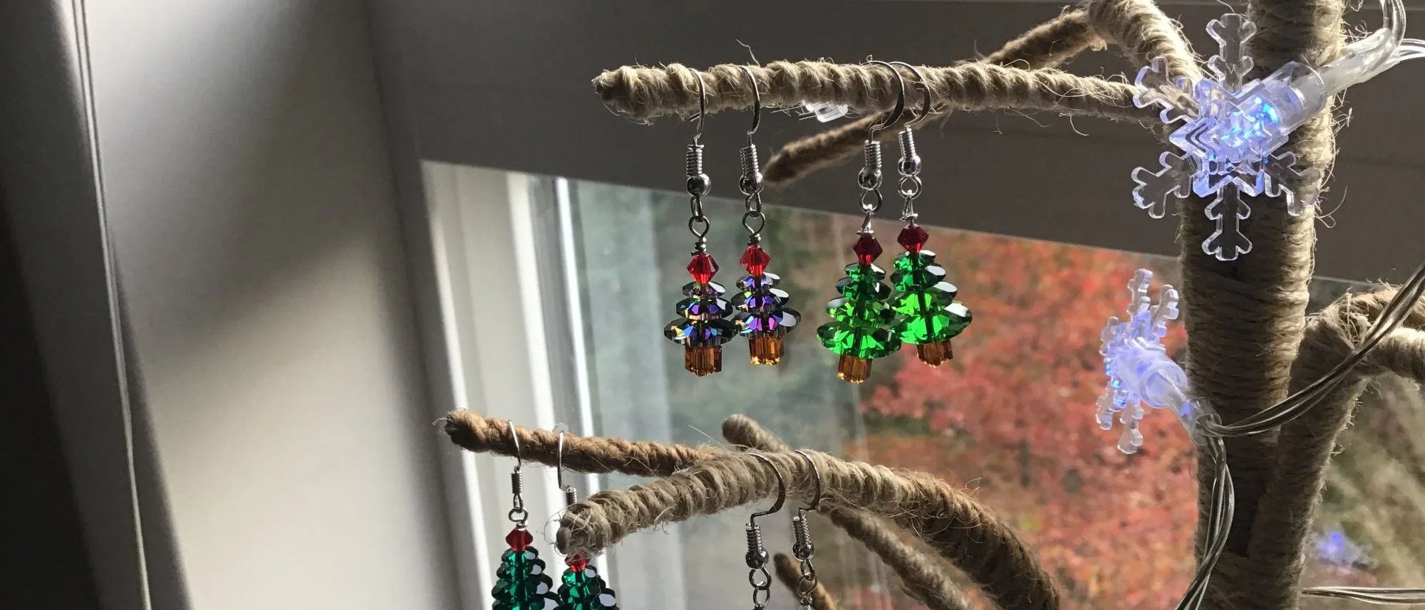 Crystal Christmas tree earrings hanging from a twine wrapped branch, green and iridescent trees topped with red beads, autumn leaves through the window behind