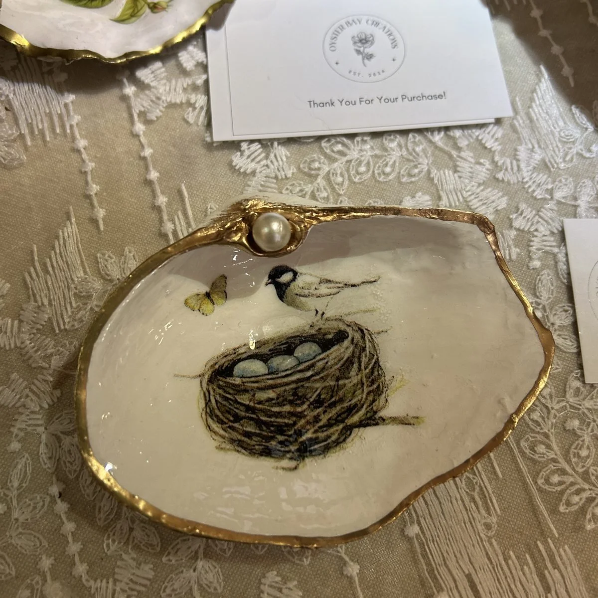 A shell dish decorated with a bird beside its nest of pale blue eggs and a yellow butterfly, gold rimmed with a pearl at the hinge
