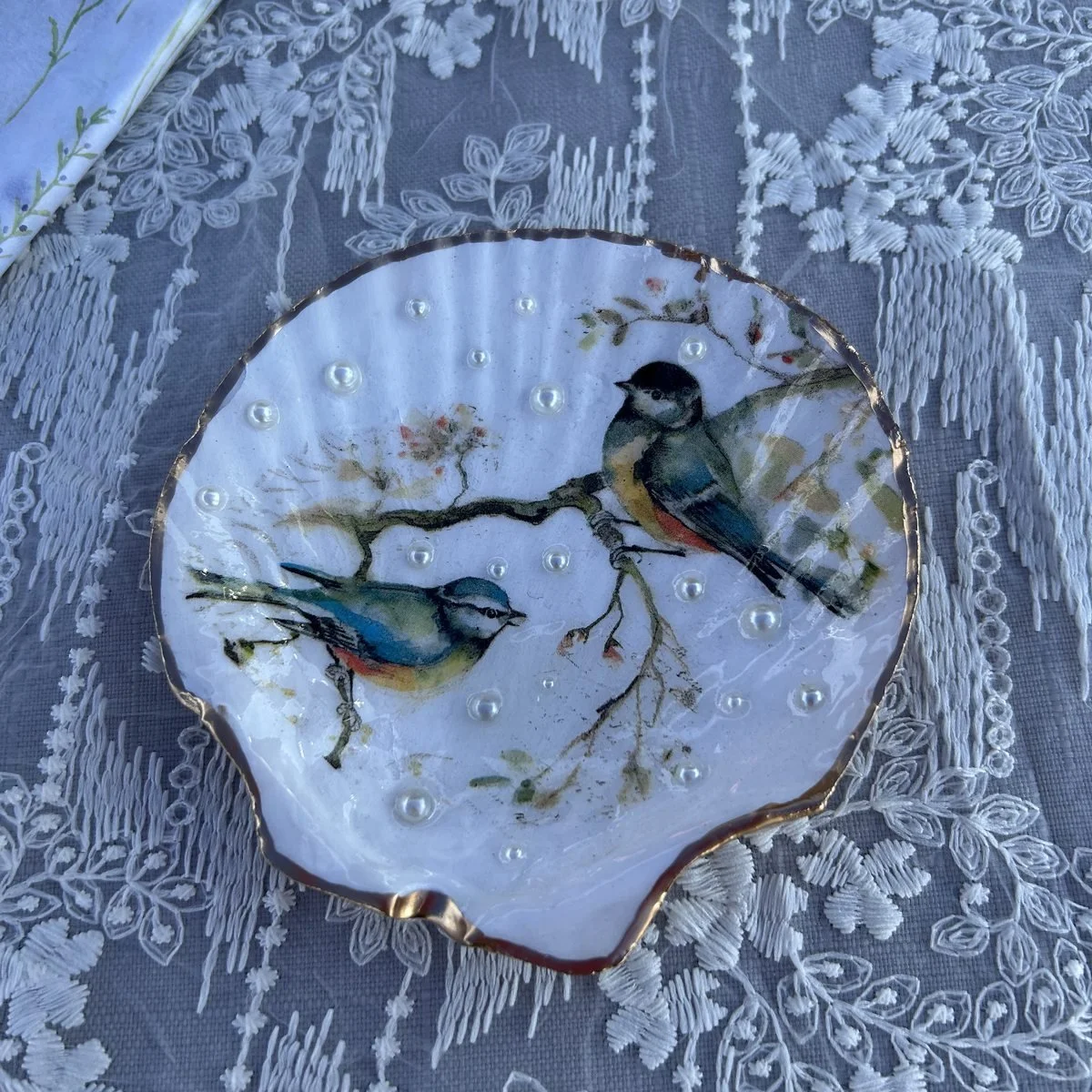 A scallop shell dish painted with two blue tits on a branch, dotted with small pearls and edged in gold