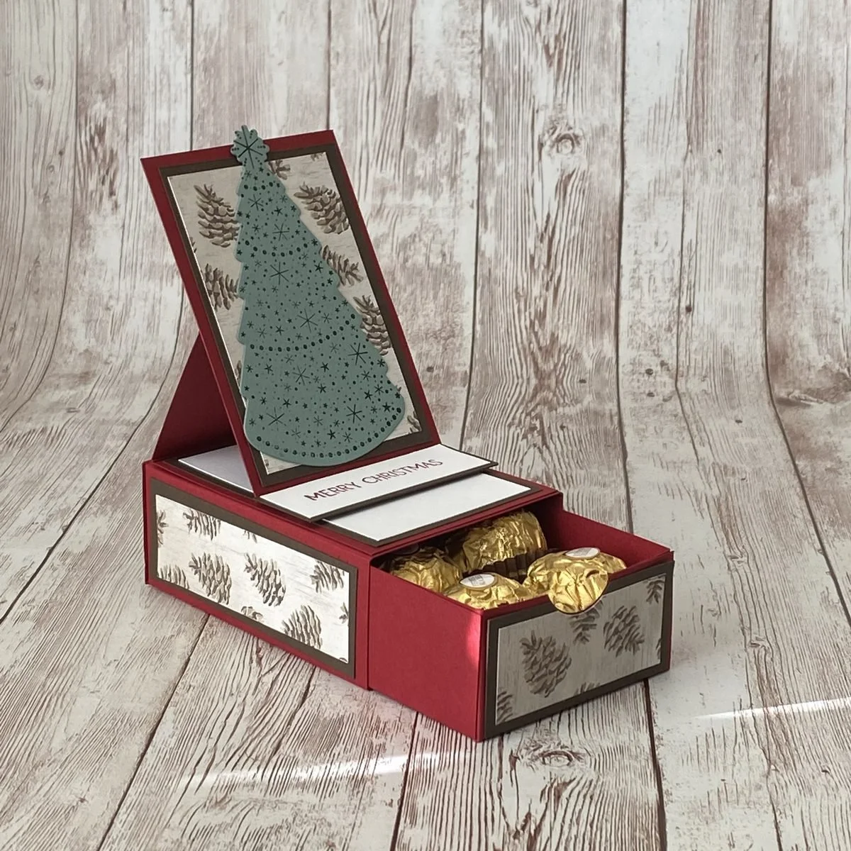 A red papercraft treat box opened to show chocolates in a drawer, with a pine cone patterned lid, a stamped Christmas tree and a Merry Christmas card tucked inside