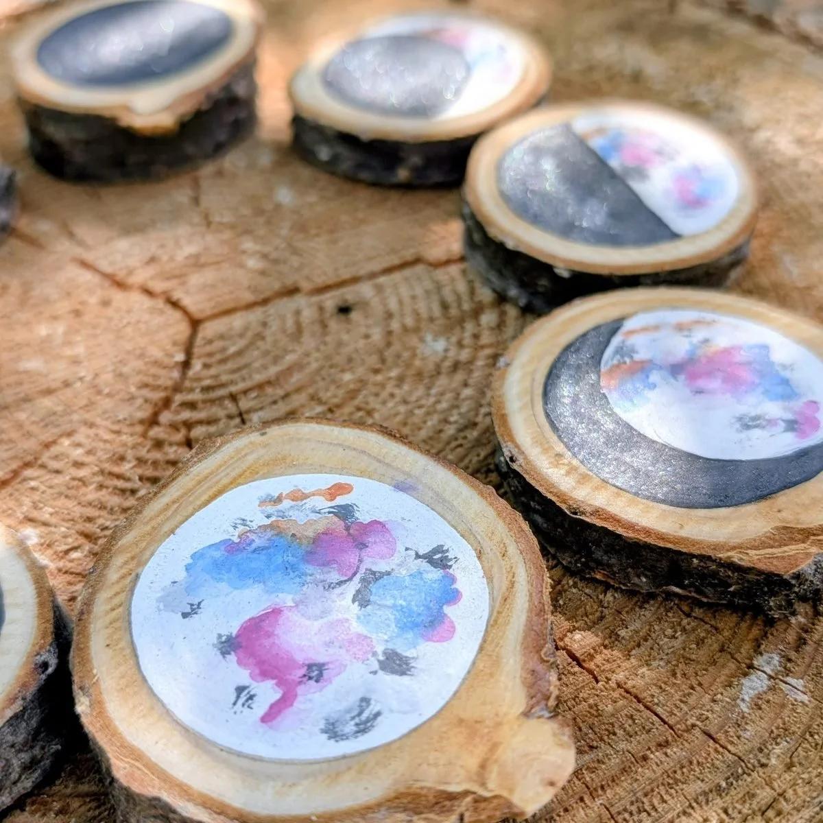 Wood slices painted with the phases of the moon in watercolour blues, pinks and silver, arranged on a tree stump