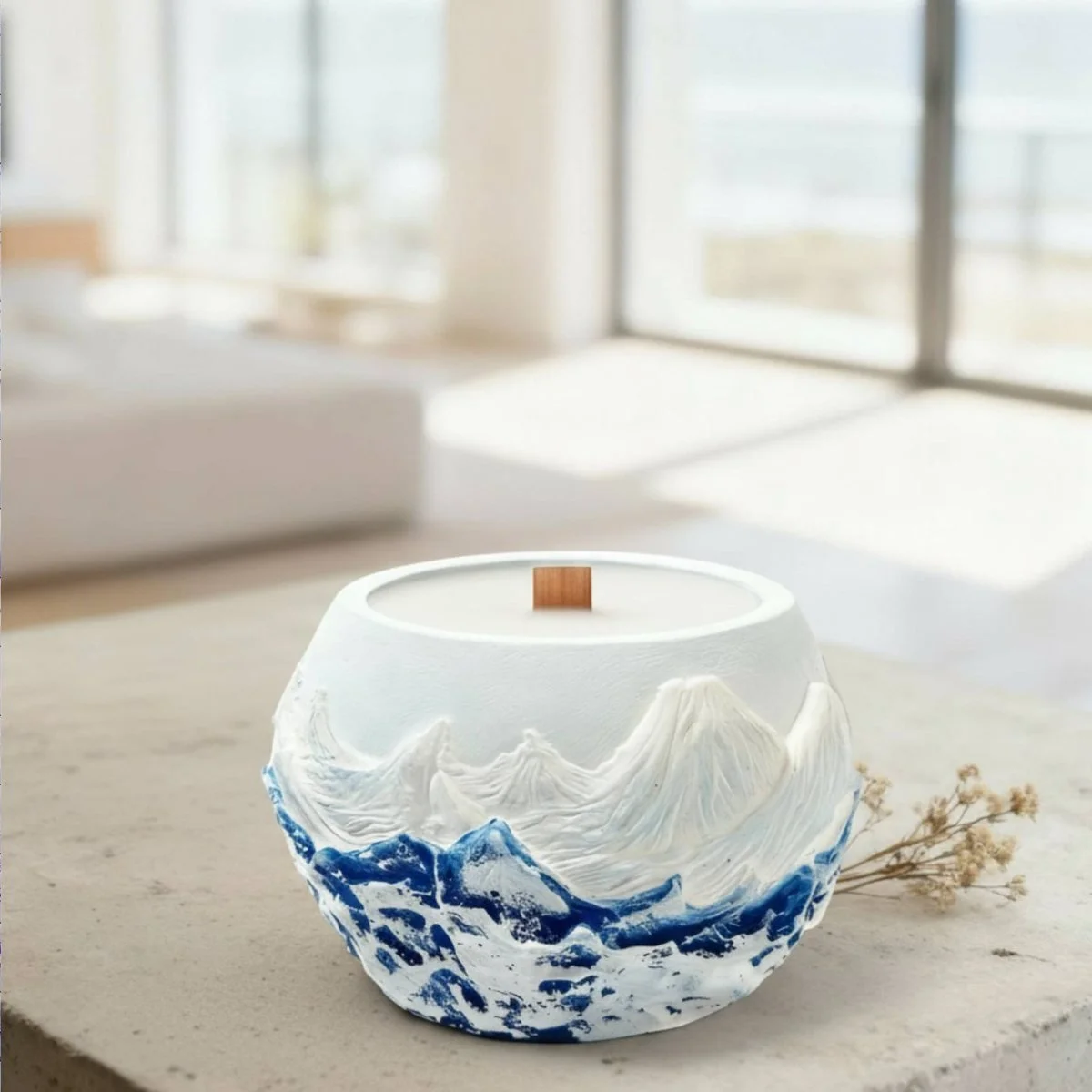 A round candle vessel carved with snowy peaks above a band of deep blue, standing on a concrete ledge