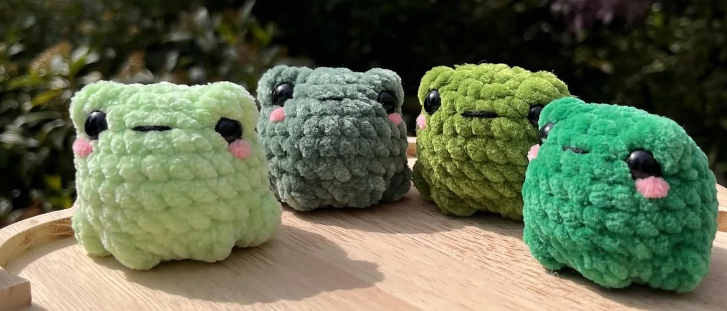 Four chunky crochet frogs in different shades of green, sitting in a row on a wooden tray with pink cheeks and black eyes