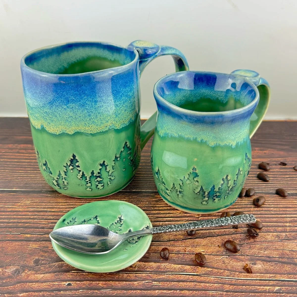 Two hand built mugs glazed blue at the rim fading to green, with a treeline of firs around the base, beside a small spoon rest and scattered coffee beans
