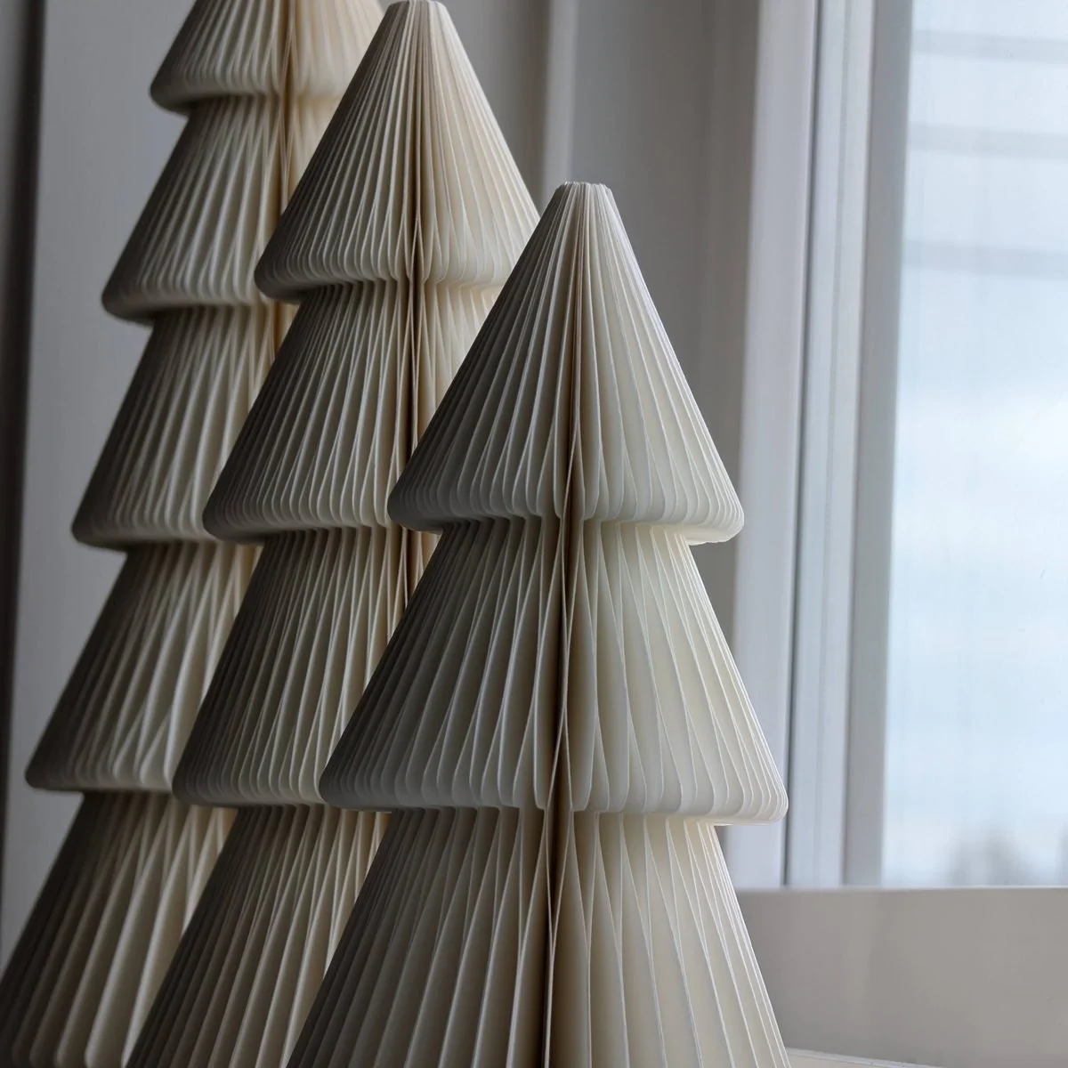 Three folded paper Christmas trees in cream and pale grey standing on a windowsill in soft daylight