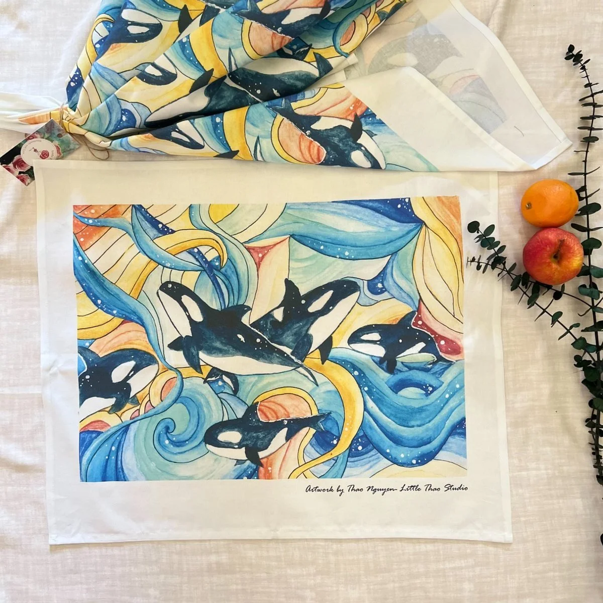 A cotton tea towel printed with a watercolour of orcas turning through blue and gold waves, laid out beside a second one folded and tied