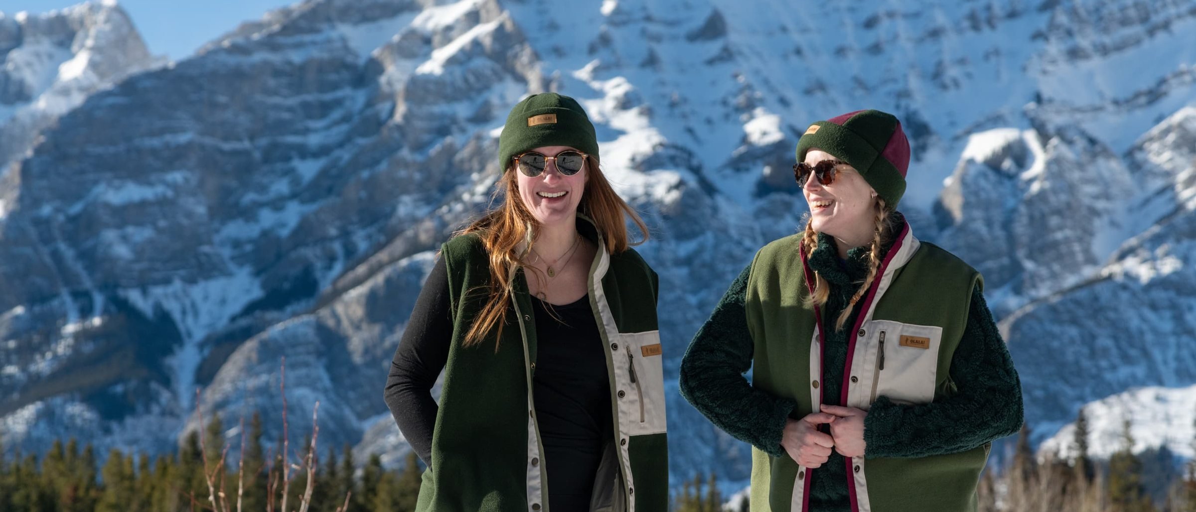 Two women laughing in green fleece vests and beanies, a snow covered mountain filling the frame behind them