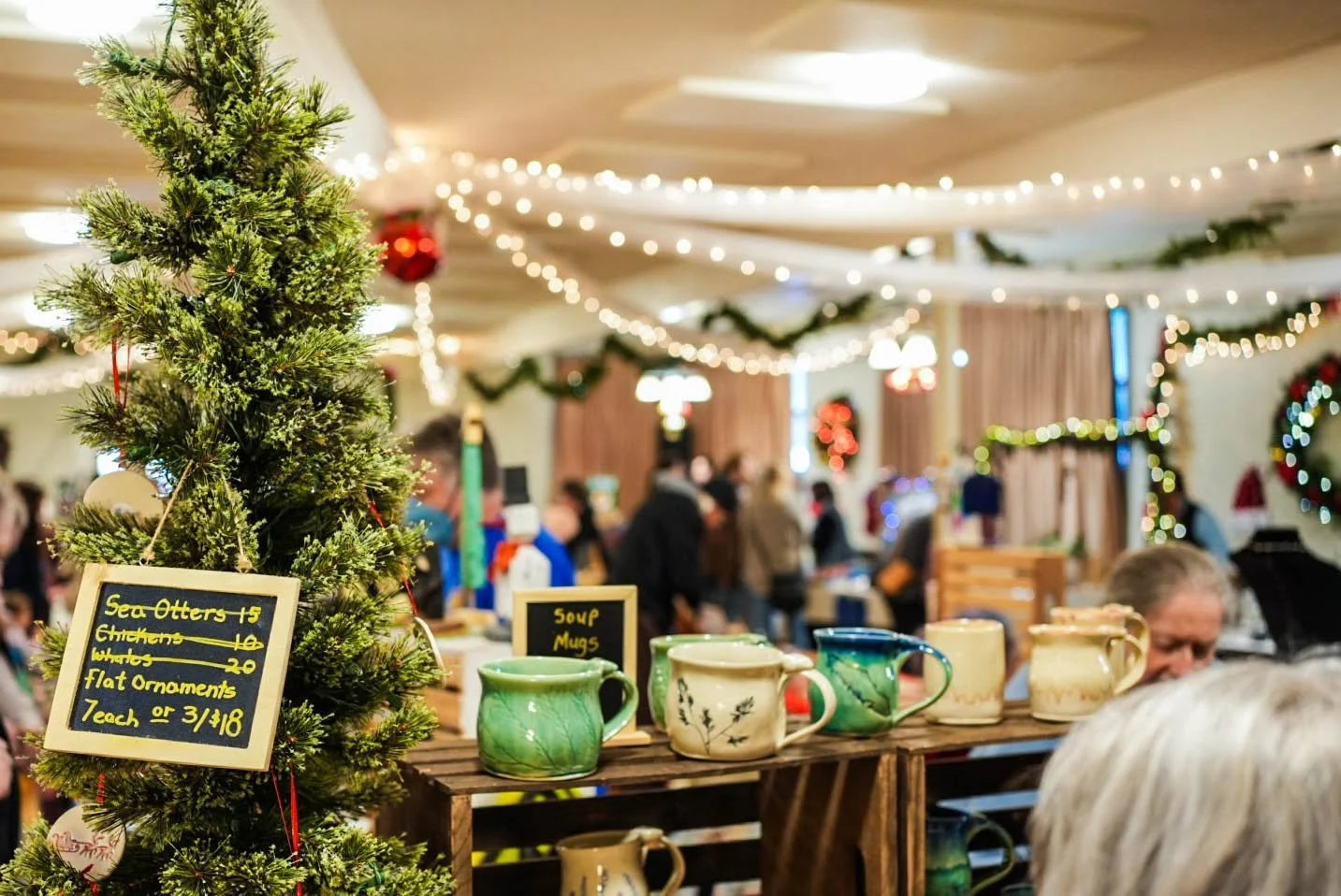 A small Christmas tree and hand-painted mugs on a vendor table, with the hall lit by strings of lights behind