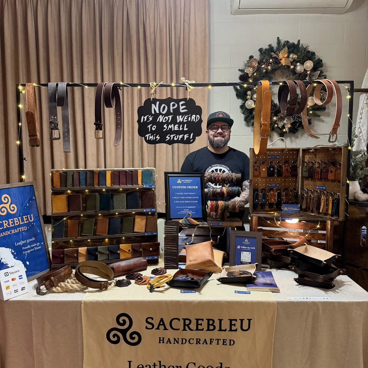 William behind his market stall, belts hanging on a rail and wallets laid out below a banner reading Sacrebleu Handcrafted, Leather Goods