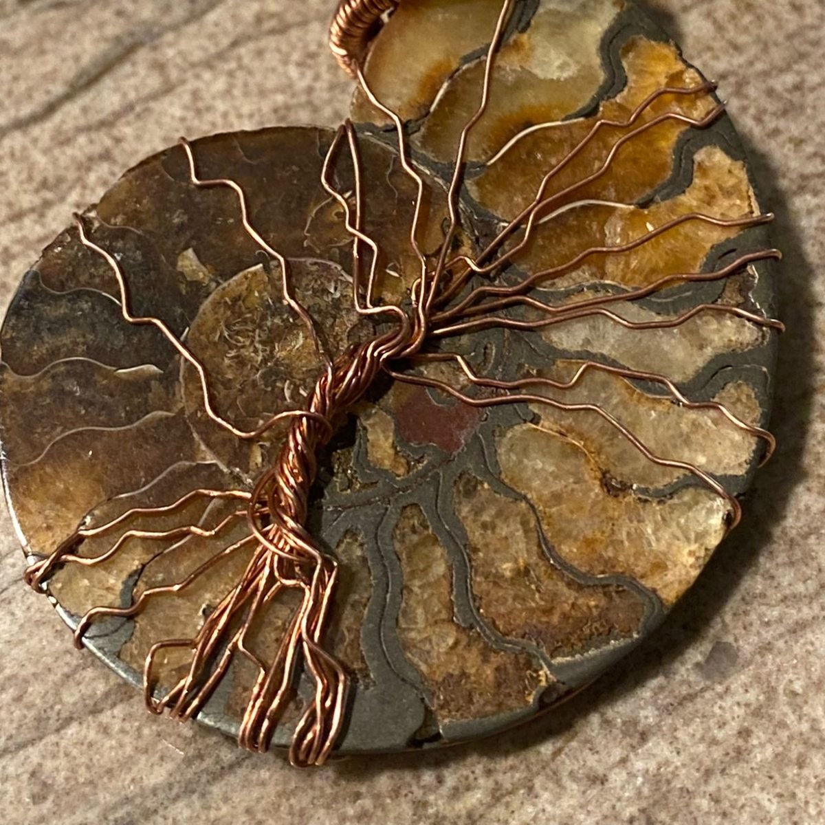 A polished ammonite fossil pendant in amber and grey, with a tree wound across the face of it in fine copper wire, hung on a copper chain