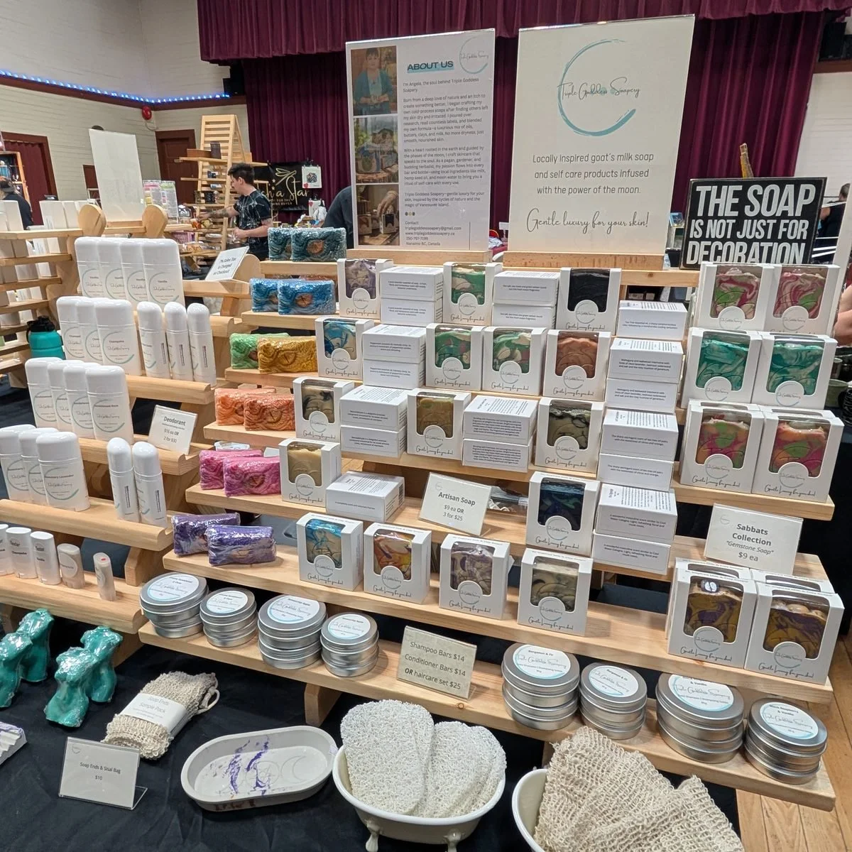 The Triple Goddess Soapery stall, boxed soaps and tins on tiered wooden shelves beneath the shop banners