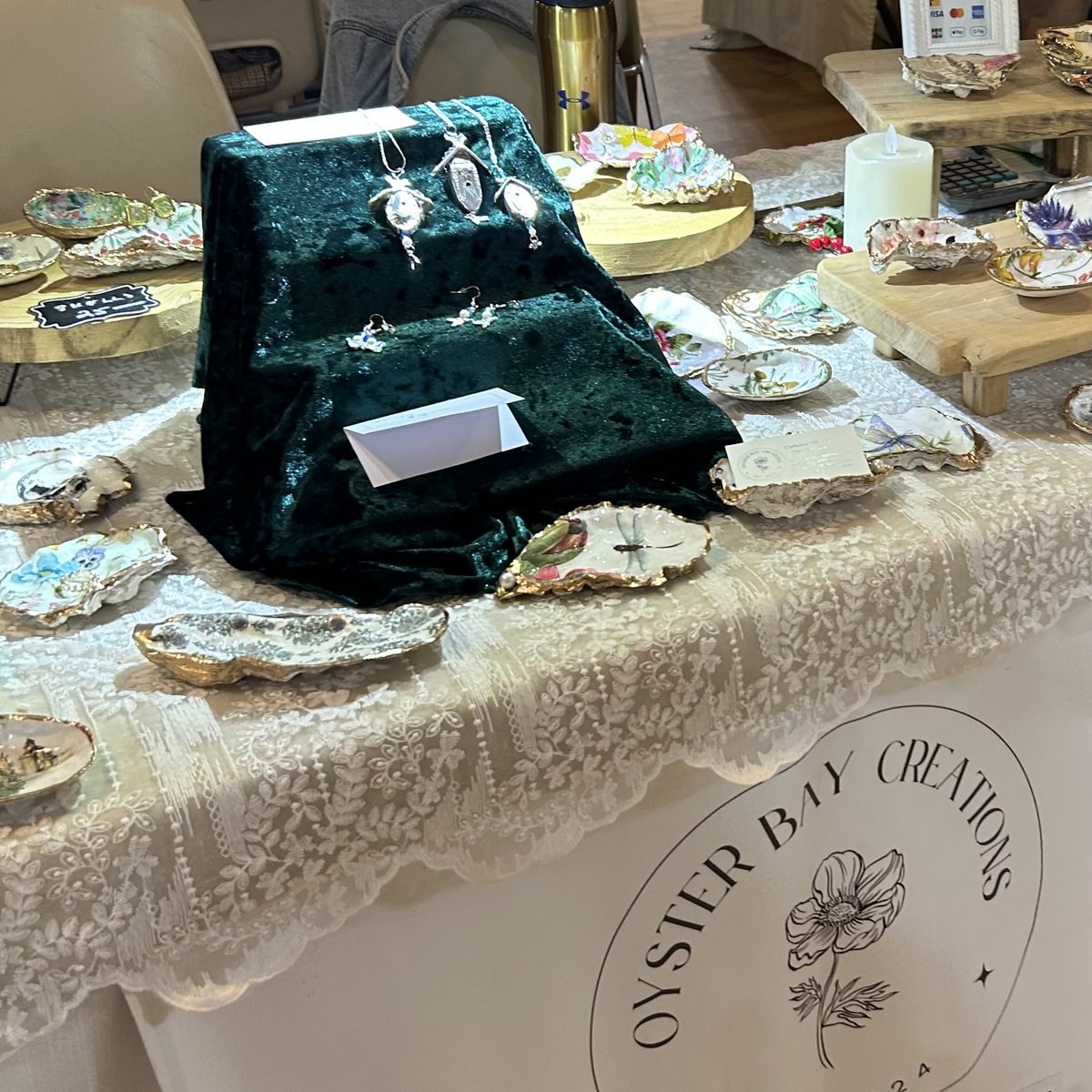 Her market table under a lace cloth, shell dishes spread across it and pendants displayed on a green velvet stand, with the Oyster Bay Creations banner below