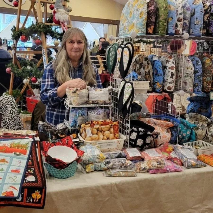 Marsha standing behind her table at a Christmas market, shelves of quilted covers and neck wraps behind her and an advent calendar wall hanging to one side