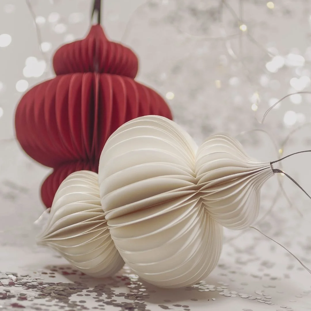 Two cream folded paper ornaments beside a deep red one, on a scatter of silver confetti with fairy lights behind