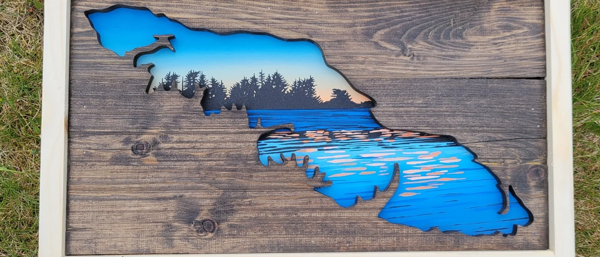 A framed panel of dark reclaimed boards with the shape of Vancouver Island cut away, revealing a painted scene of a treeline against a sunset sky above open water