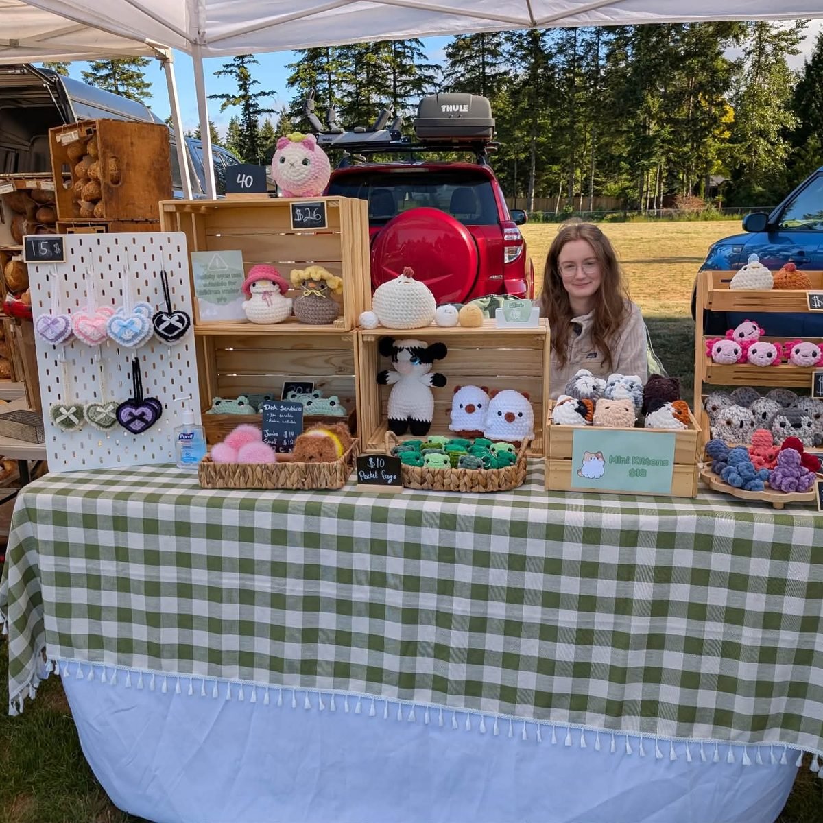 Ellie behind her market stall, a gingham covered table with crates of crochet plushies, keychains and heart charms on a pegboard