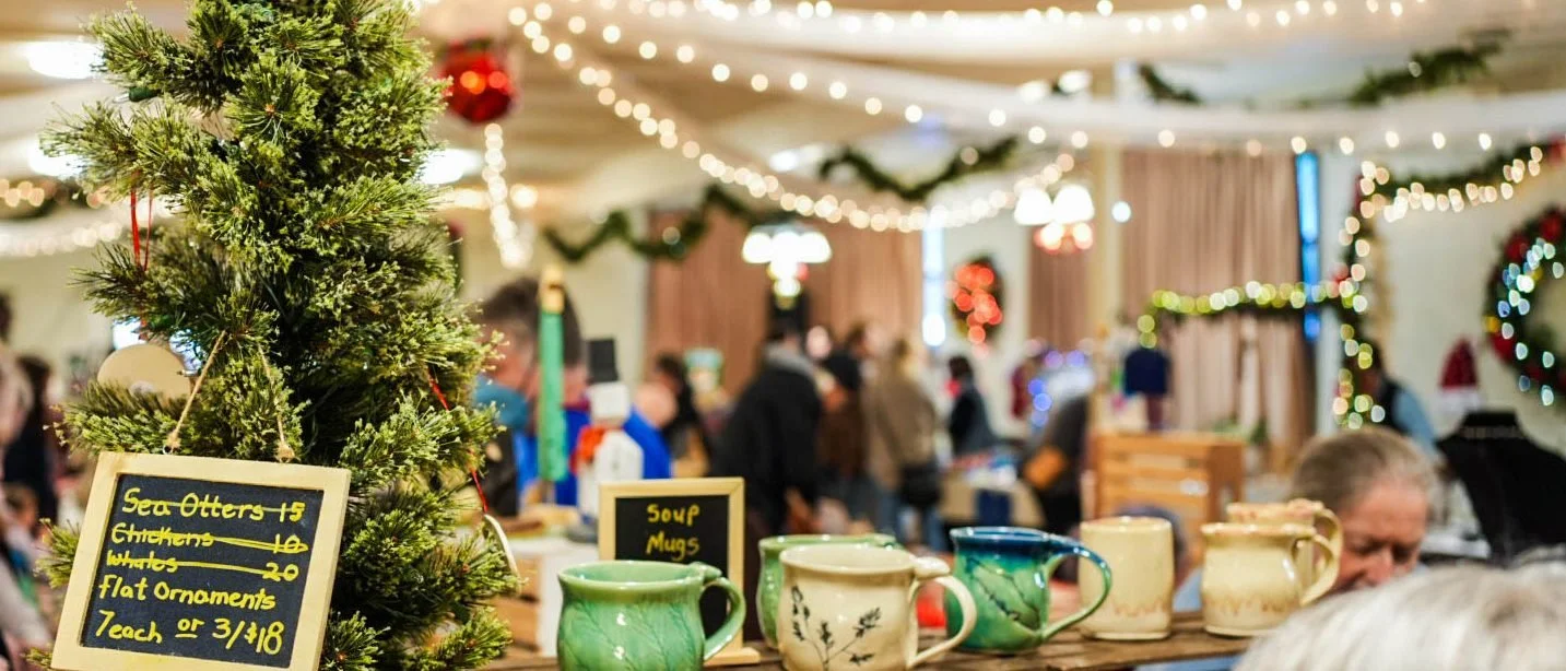 The FaeGarten Clay table at the market, hand built mugs lined up on a wooden crate beside a small tree hung with ornaments, with string lights across the hall behind