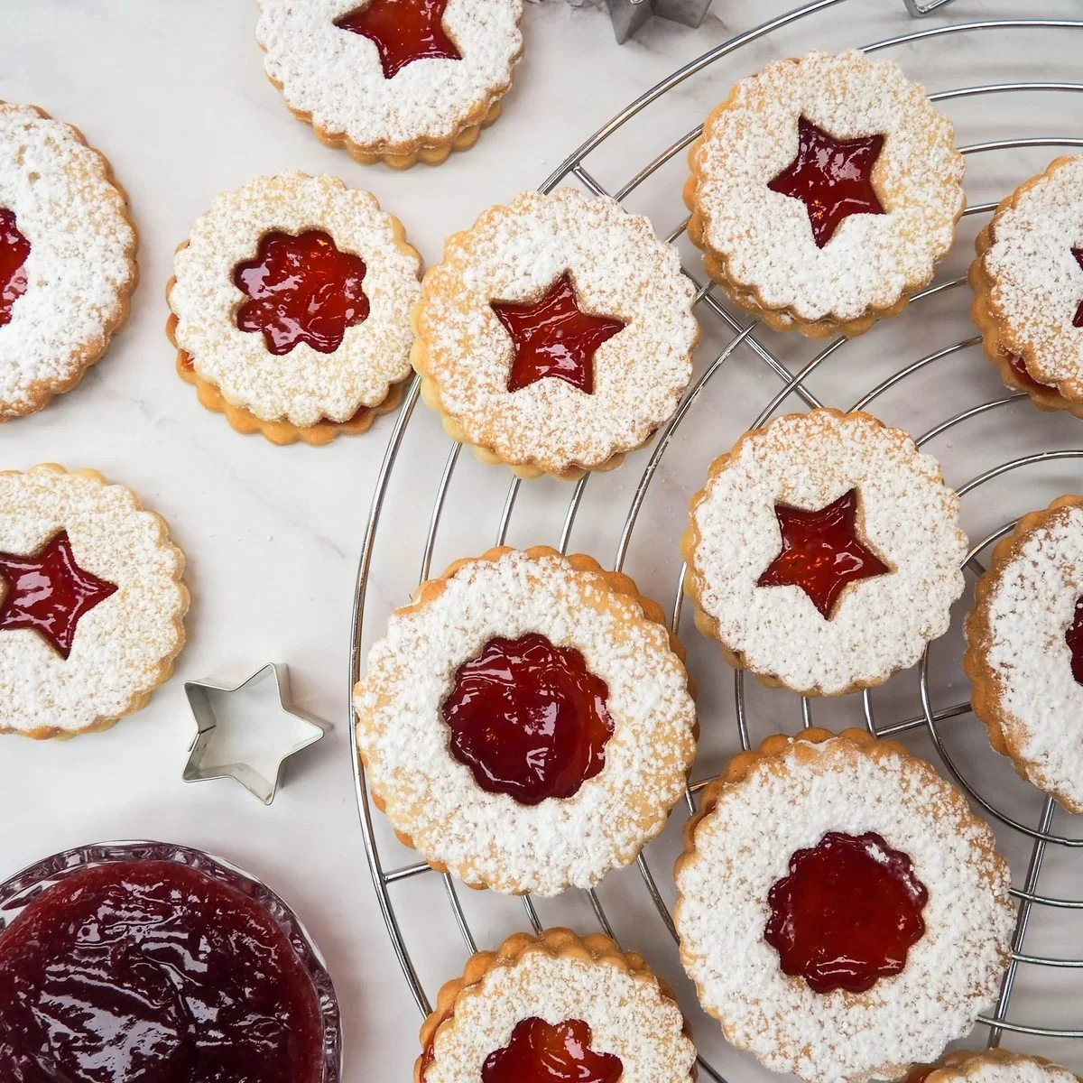 Linzer cookies dusted with icing sugar, each cut with a star or flower to show the red jam beneath, cooling on a wire rack