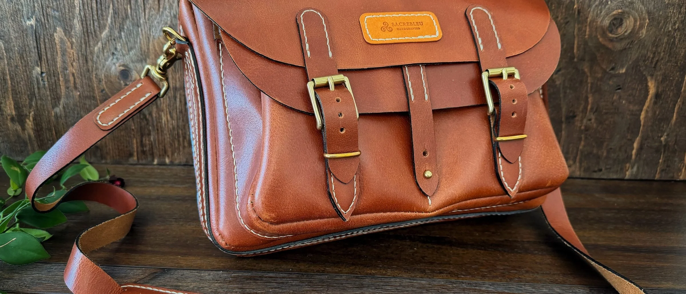 A tan leather satchel with brass buckles and cream hand stitching, its long shoulder strap laid out in front of it on dark wood