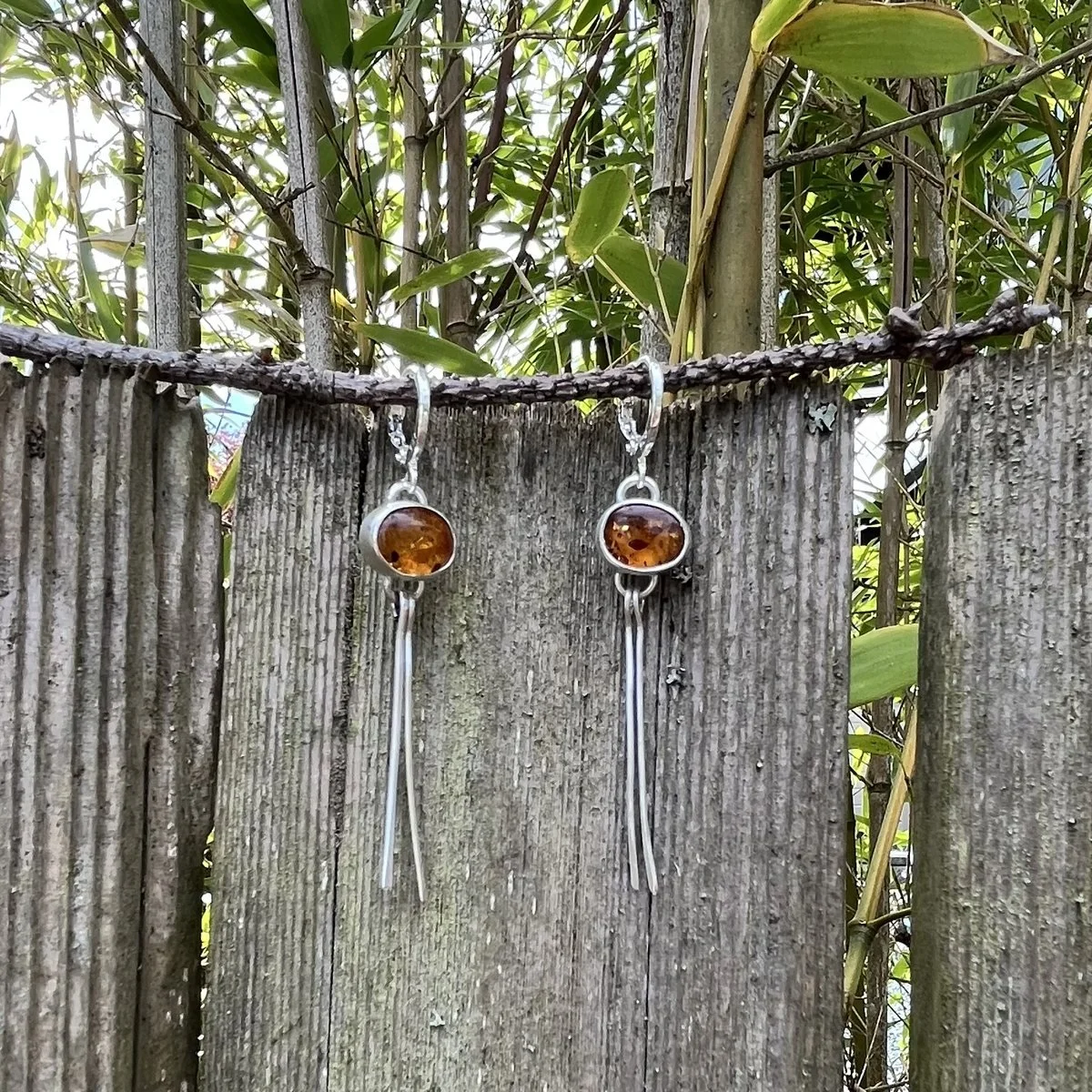 A pair of earrings hanging from a branch, each an oval amber cabochon in a silver bezel above two long hammered silver strands