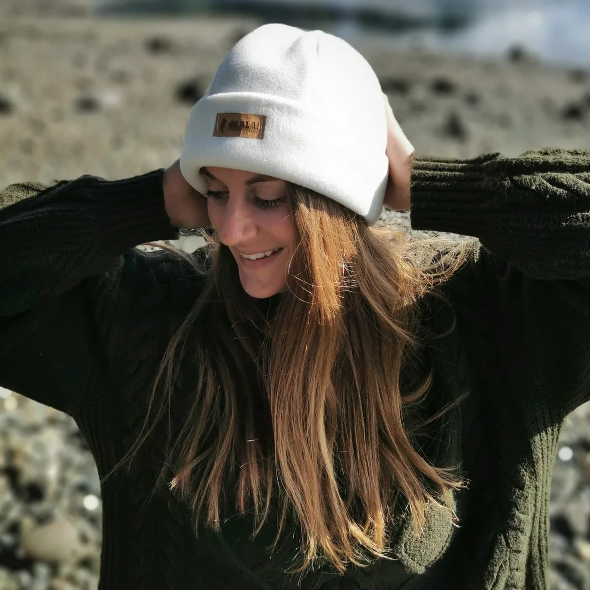 A woman on a pebble beach pulling on a cream fleece beanie, wearing a dark green cable knit sweater