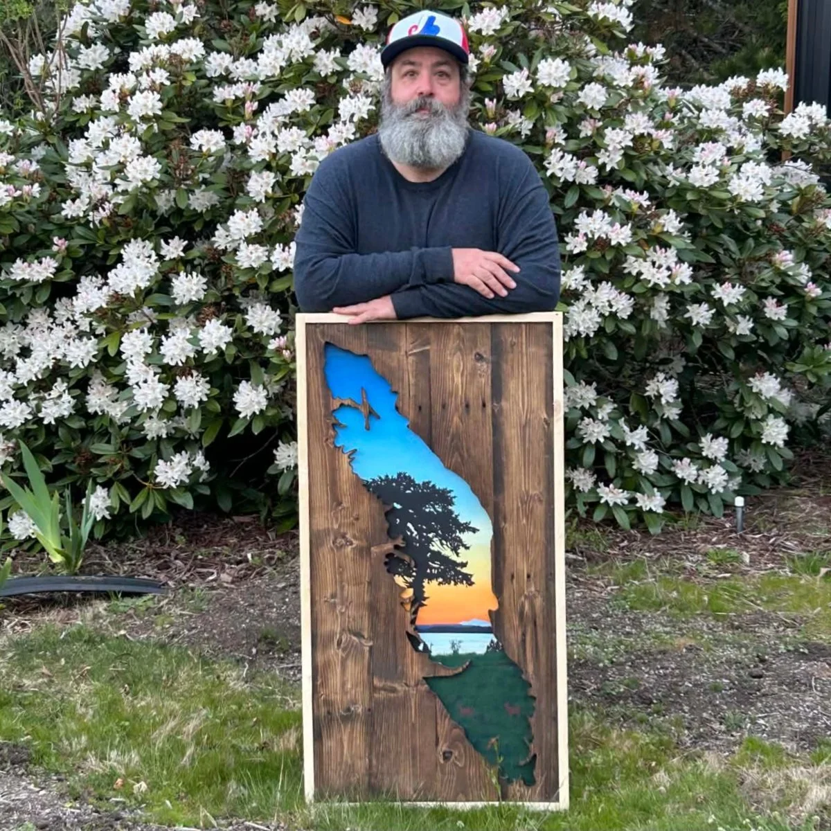 Kent standing behind one of his tall island panels in front of a flowering rhododendron, arms resting along the top of the frame