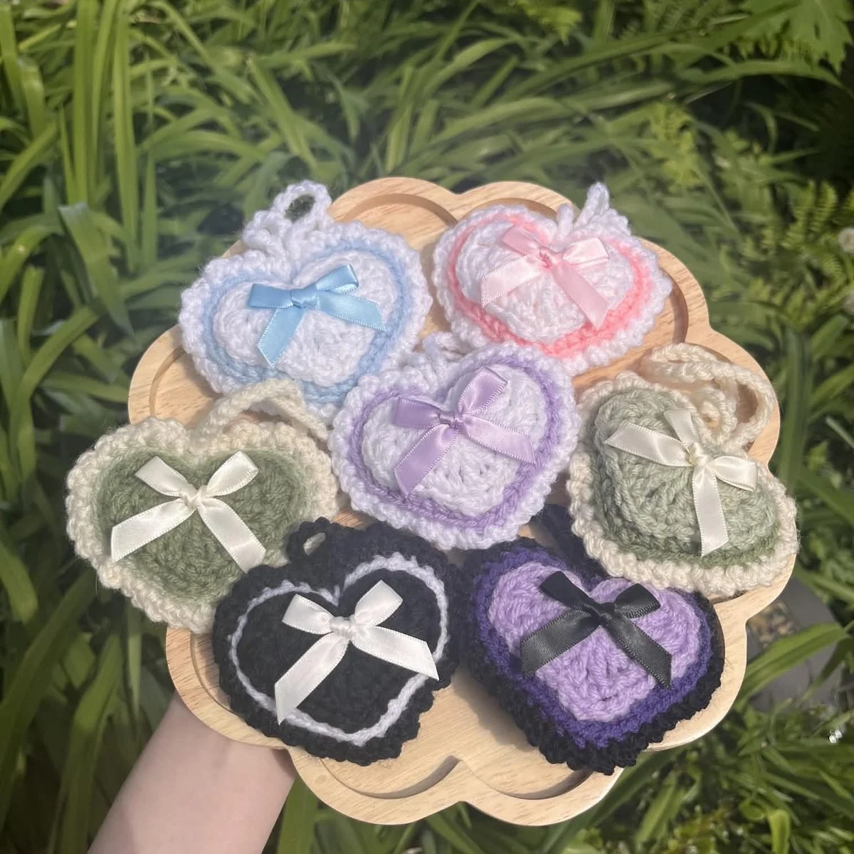 Crochet heart charms on a wooden tray in blue, pink, lilac, sage, black and purple, each finished with a satin bow