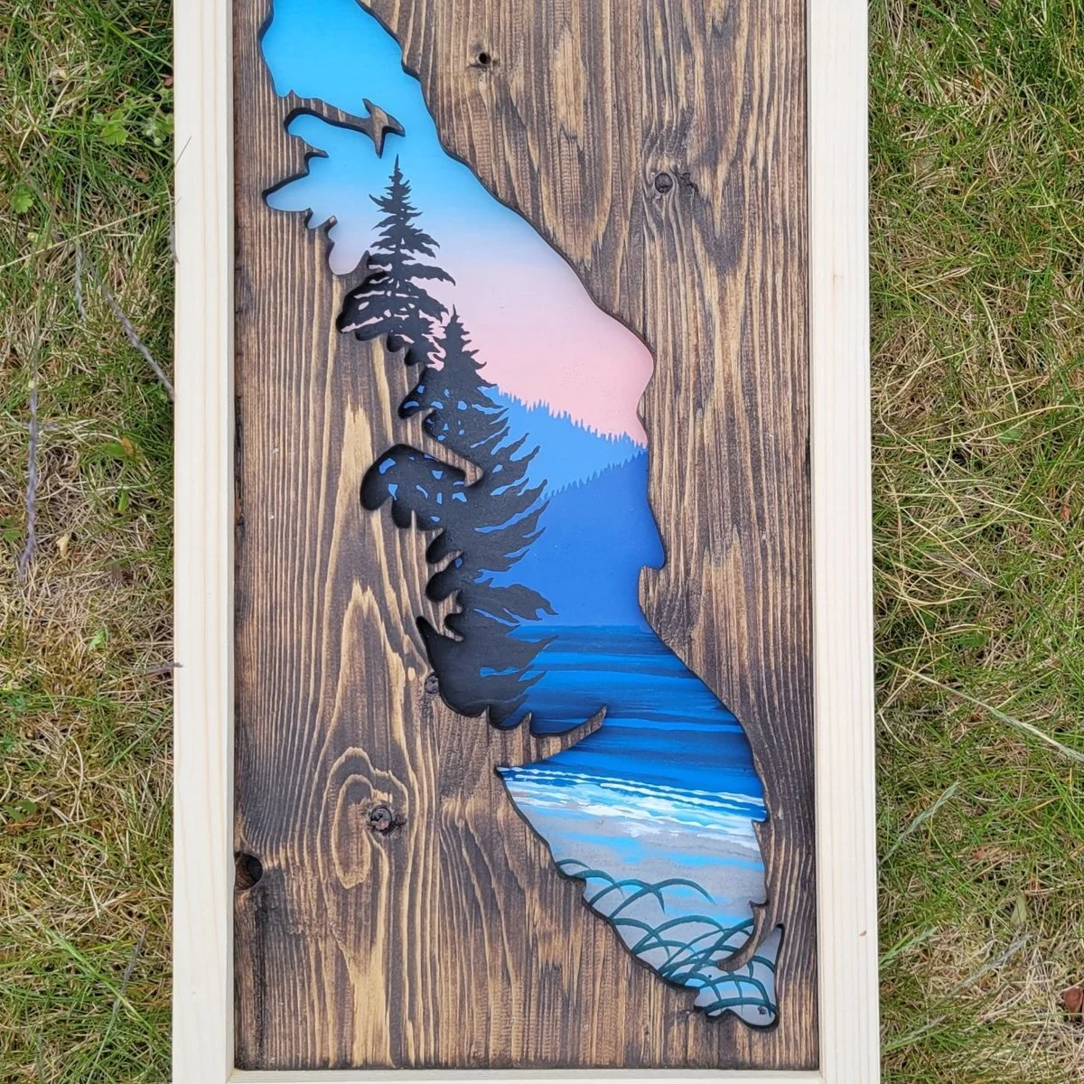 An upright cutout of Vancouver Island in reclaimed wood, the opening painted with a windswept fir above a beach and a pink and blue dusk sky