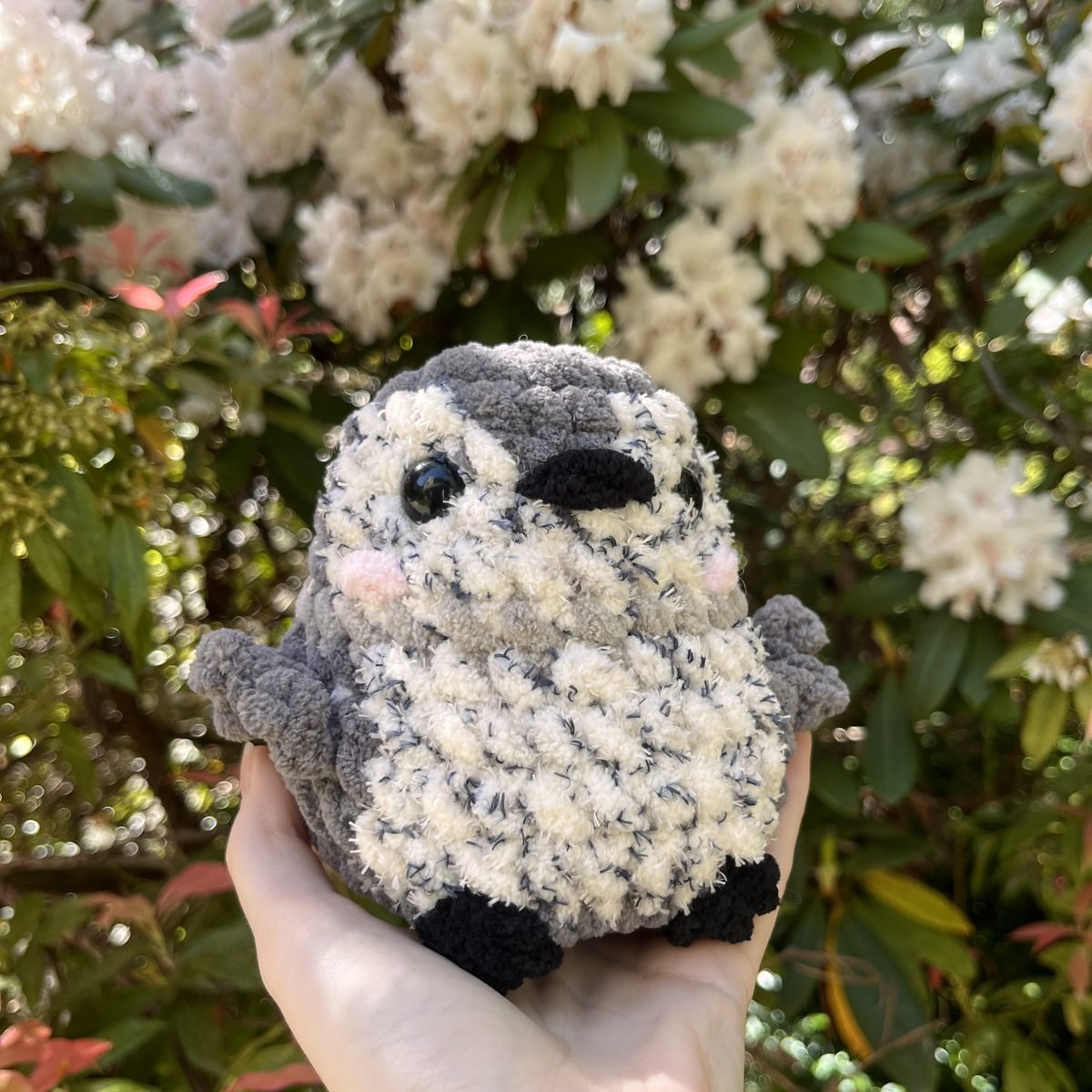 A crochet penguin plushie in speckled grey and cream held in one hand, in front of a flowering rhododendron