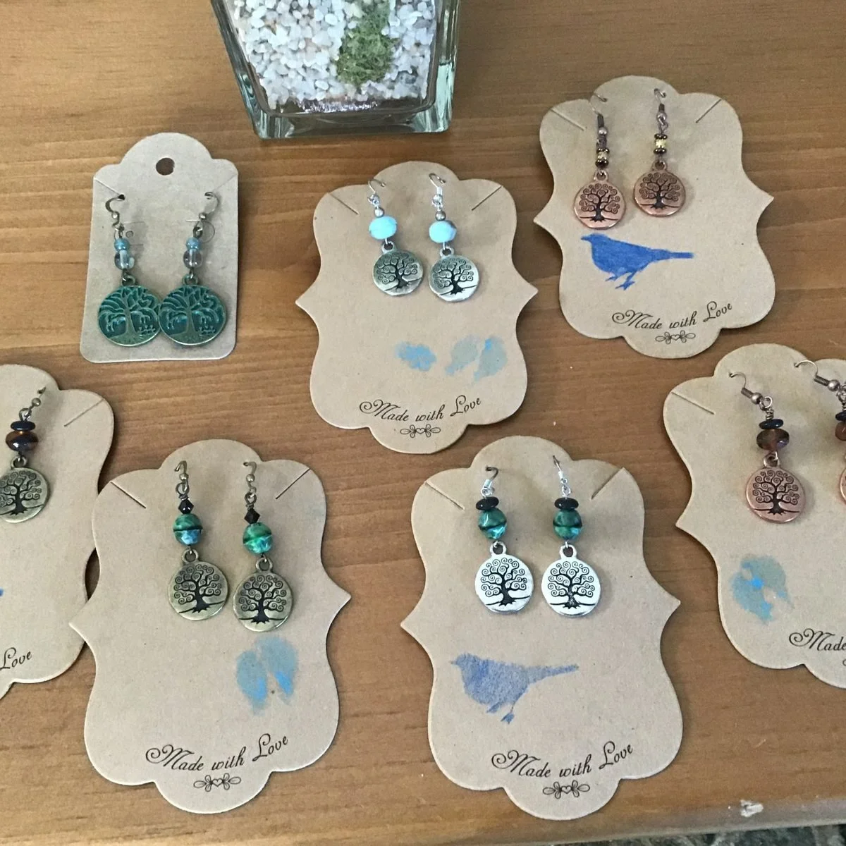 Seven pairs of tree of life earrings on kraft cards printed with a bluebird and the words made with love, in silver, bronze, copper and verdigris