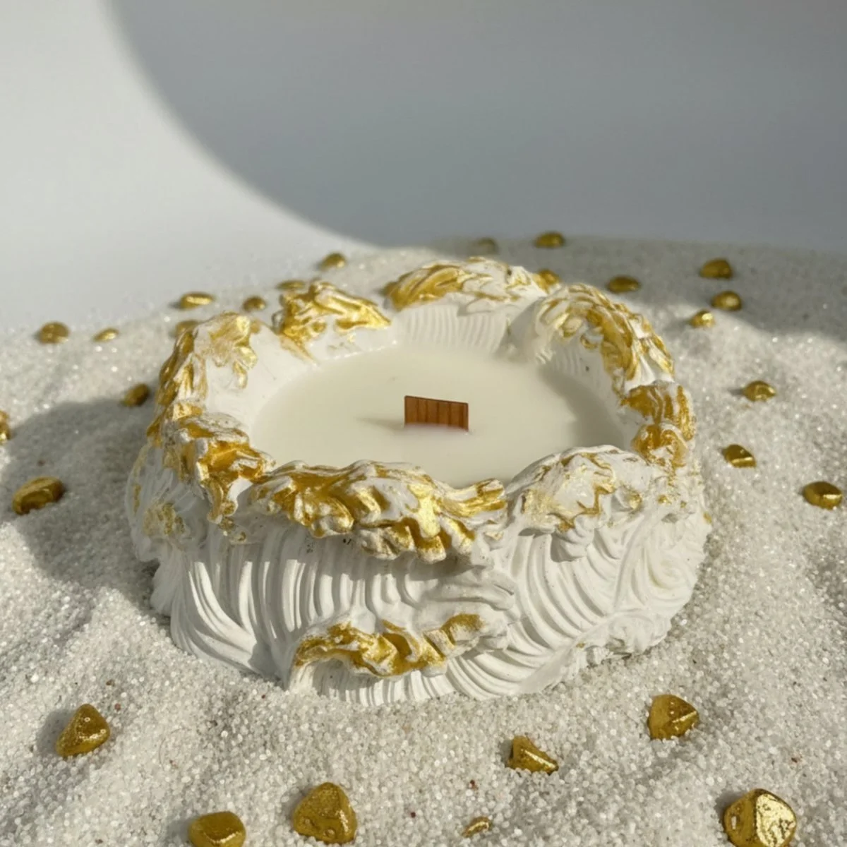 A candle vessel sculpted as a breaking wave in white with gold leaf along the crest, set in pale sand