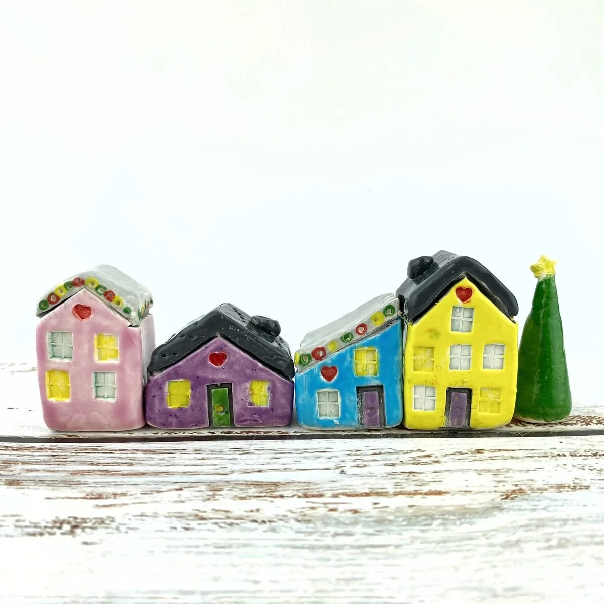 Four tiny ceramic houses in pink, purple, blue and yellow with heart details over the doors, lined up beside a small green tree