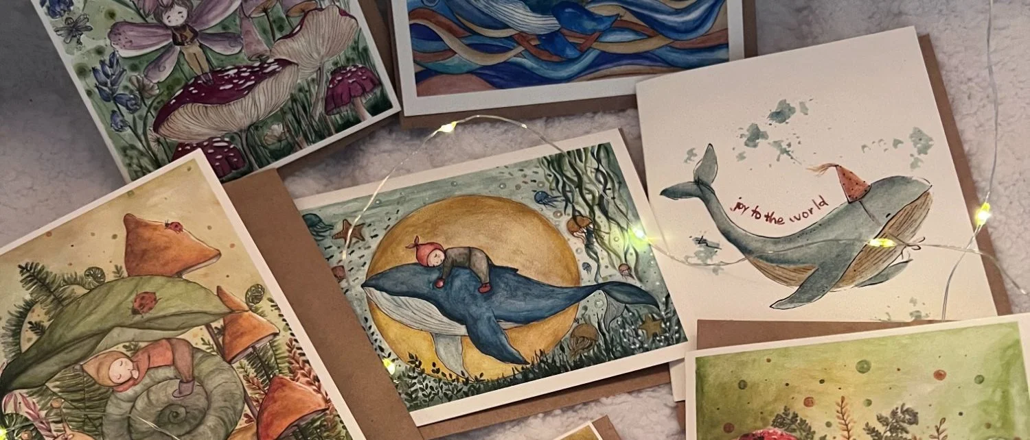 Watercolour greeting cards spread out on a white surface among fairy lights, showing whales, fairies, mushrooms and a card reading joy to the world