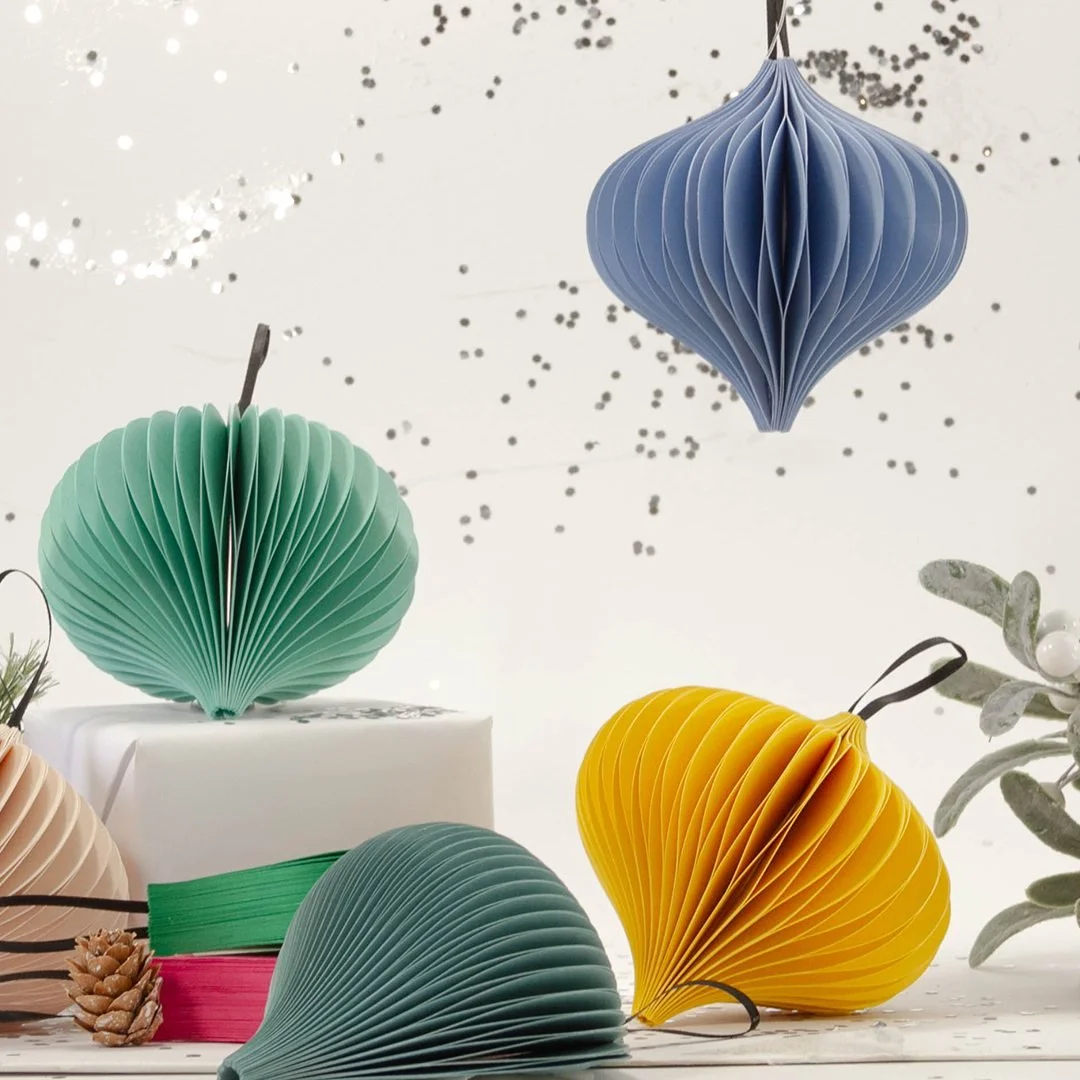Folded paper ornaments in teal, blue, yellow and blush arranged around a wrapped gift, with silver confetti falling behind