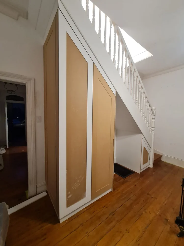 The full run in raw MDF: tall cabinet at the high end, framed doors mid-run, open nook under the low stair