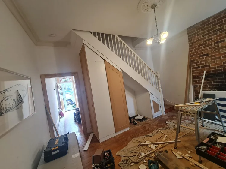 The hallway as worksite: raw MDF joinery fitted under the stair, sawhorse and tools out, ceiling rose and exposed brick beyond