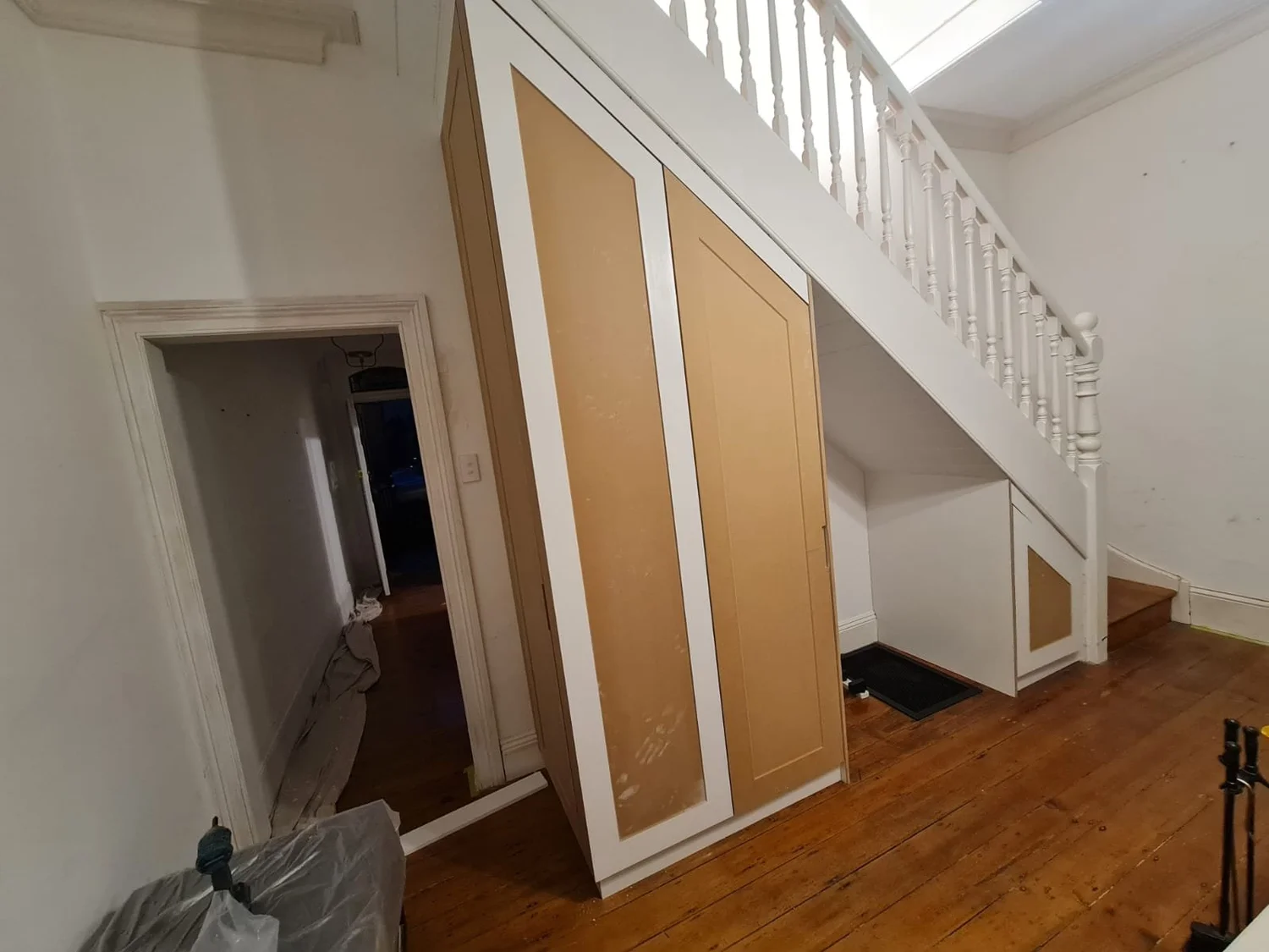 Under-stair storage mid-installation in a heritage terrace hallway: raw MDF panelled doors and a triangular end door following the line of the white staircase