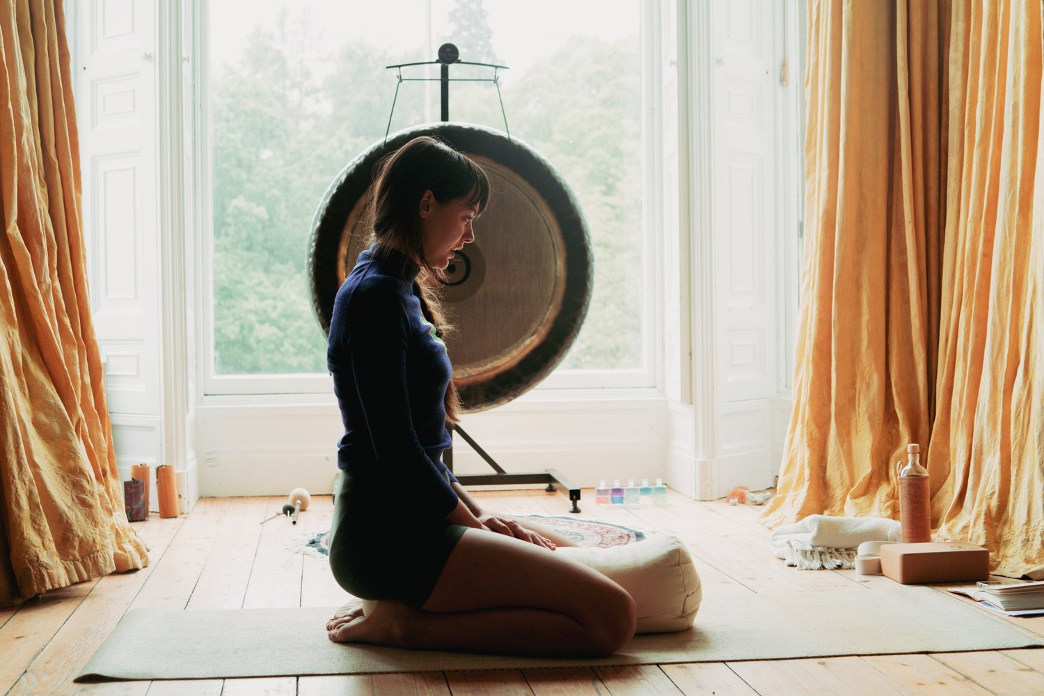 A woman kneeling on a yoga mat in front of a large gong, framed by tall windows and gold curtains.