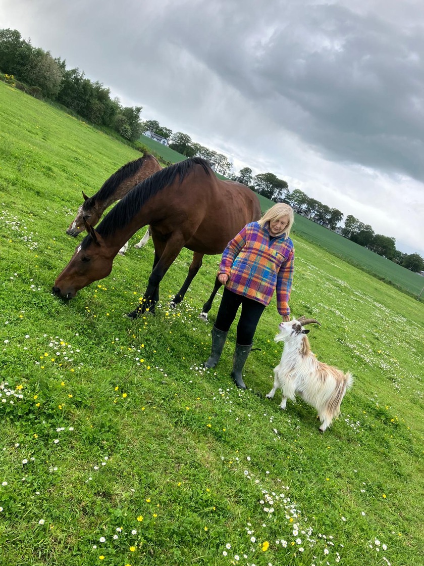 Marianne standing in a field in a bright checked shirt, surrounded by her goats with a white pony beside her.