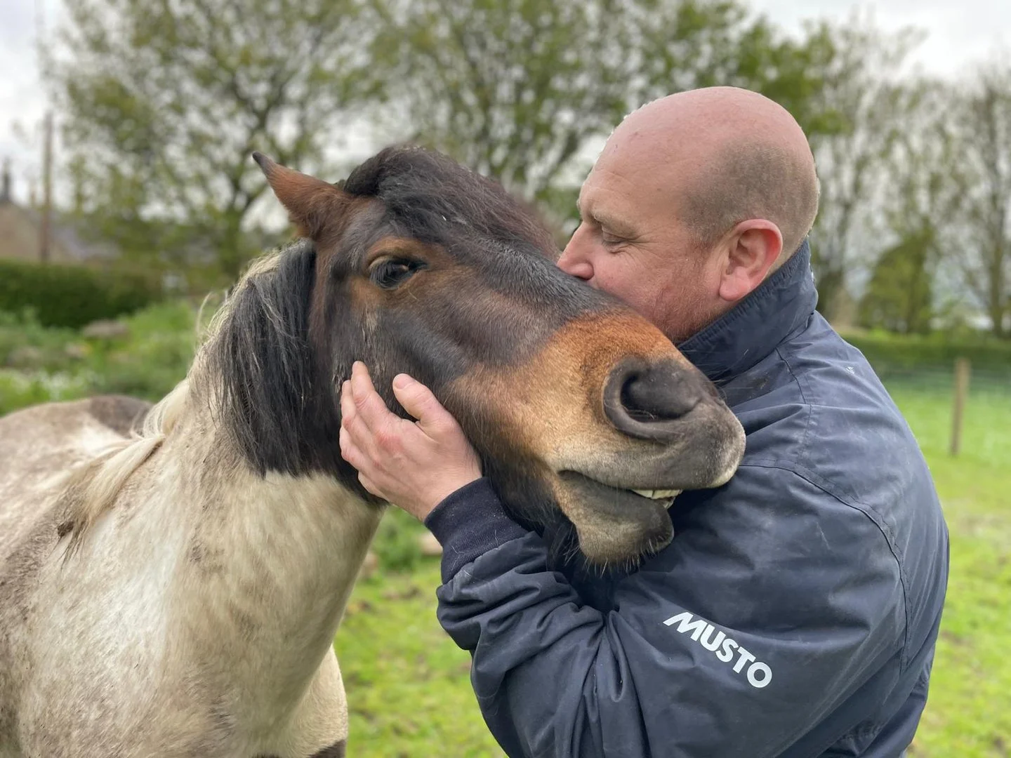 A man in a navy jacket holding the head of a roan pony close against his face, both of them still and calm.