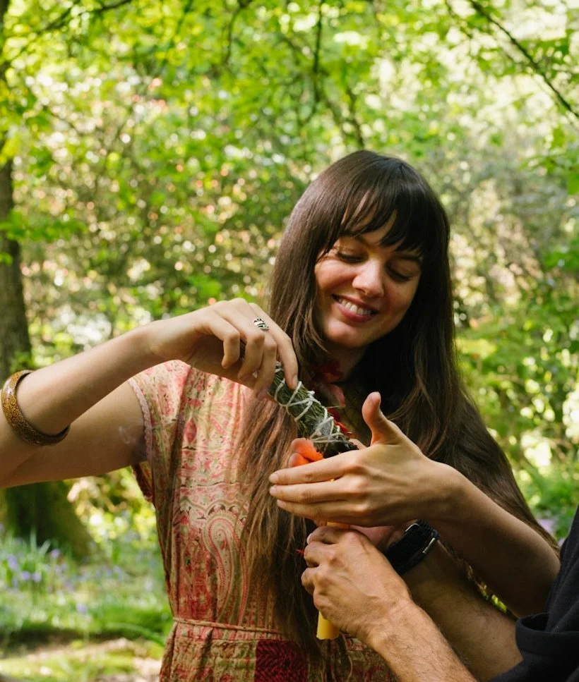 Alexandra outdoors in dappled sunlight, wearing a patterned dress and smiling as she looks down at something held in her hands.