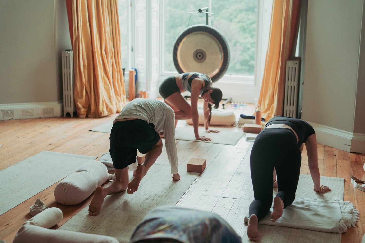 A small group practising yoga together on mats in a bright room, folding forward with a gong standing behind them.