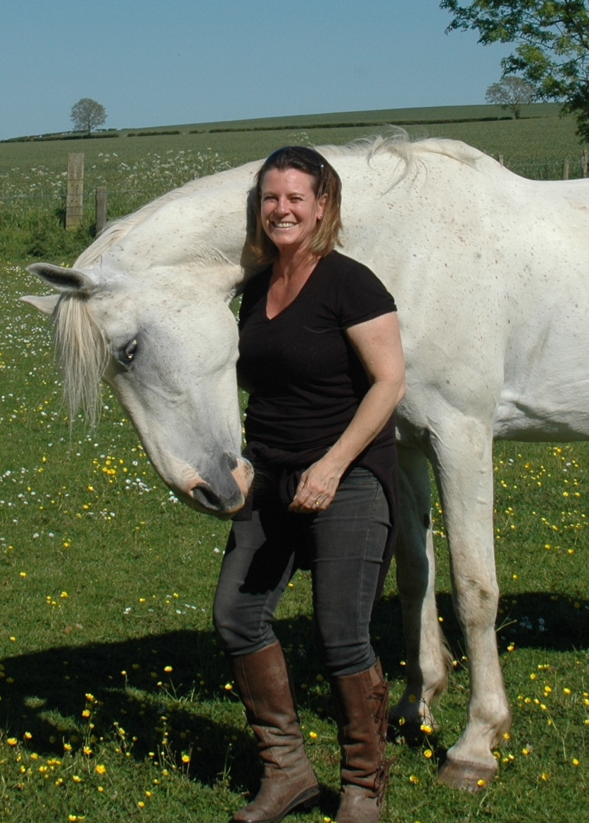 Sue standing in a buttercup meadow with her arm resting over the neck of a grey horse, both facing the camera.