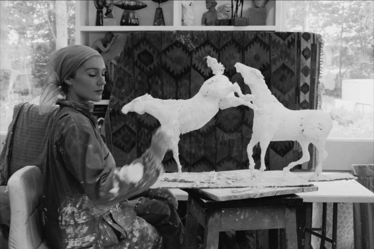 Black and white photograph of Jeanie at work in her studio, shaping a plaster horse maquette on a bench surrounded by finished pieces.