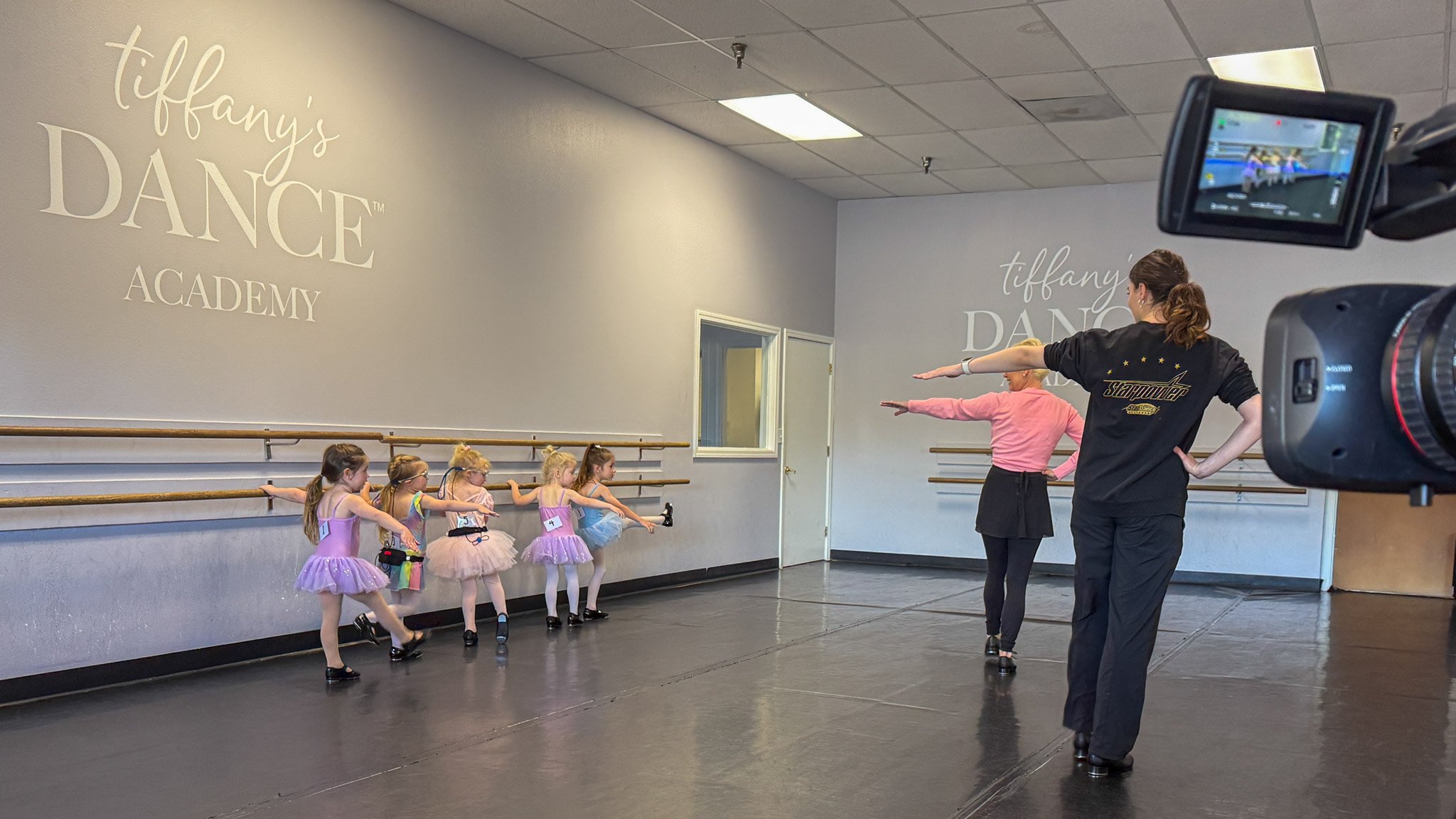 Young dancers wearing eye-tracking glasses at the barre during the study class at Tiffany's Dance Academy, with an instructor demonstrating and a camera recording