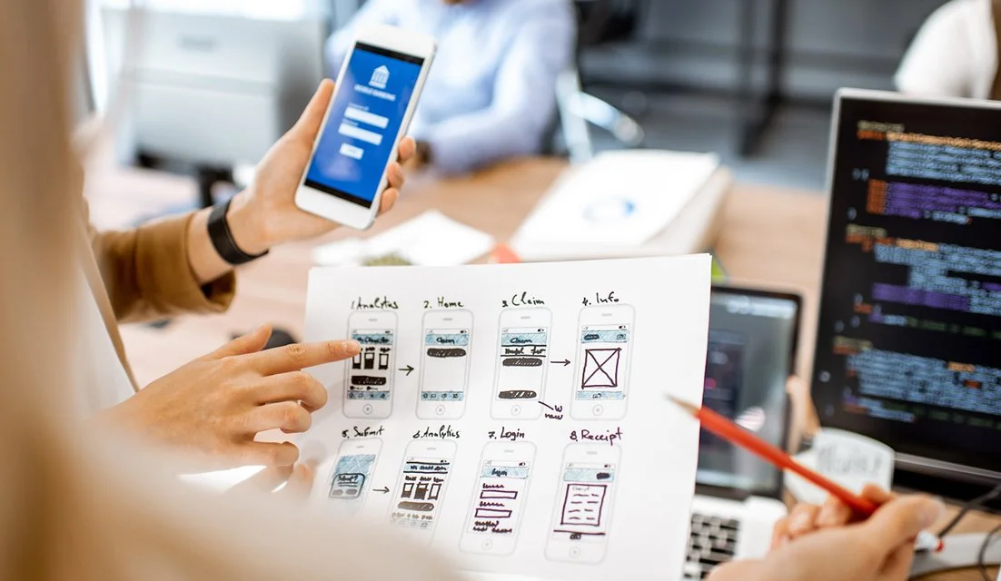A group of people collaborating around a table, with one person holding a smartphone displaying a login screen, and a paper with hand-drawn app interface sketches, in a creative office setting.